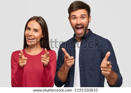 Horizontal shot of happy friends point fingers at you, gesture indoor, make choice, have positive expressions, isolated over white background. Joyful brunette woman and her lover model indoor