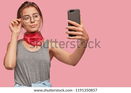 Satisfied European woman takes images on modern cell phone, shares in blog, wears transparent glasses for good vision, dressed casually, poses against pink background with blank space for your text