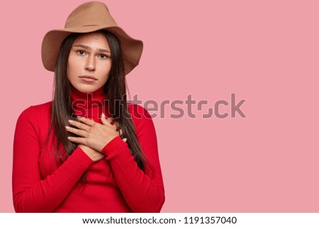 Sad serious freckled woman with unhappy expression, keeps hands on chest, asks to listen her and forgive bad actions, wears casual clothes poses over pink background with free space for your promotion