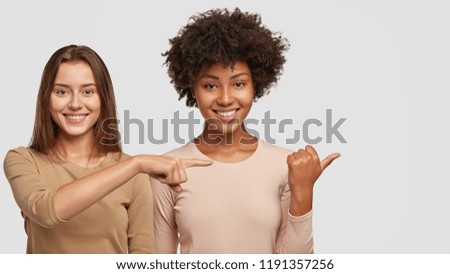 Photo of satisfied interracial young women point aside with joyful expressions, show nice place to visit, have tender smiles on faces, stand closely to each other, isolated over white background.