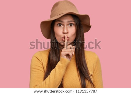 Hush, please! Beautiful young brunette woman keeps index finger over lips, has surprised expression, demands silence, isolated over pink background. Young teacher asks pupils not make noise.