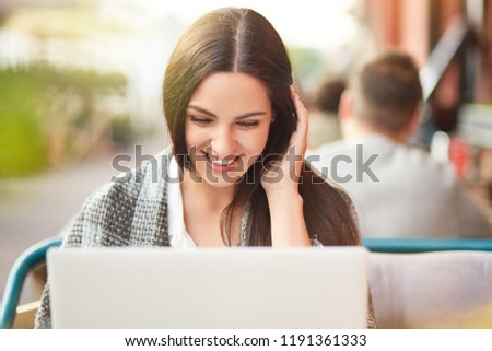 Photo of cheerful satisfied Caucasian woman looks positively at screen of laptop computer, has coverlet, reads funny stories on website, poses in outdoor cafe. People, leisure, rest concept.