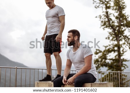 Fit sporty men relax after outdoor training session, sitting on crossfit blocks on the peak of mountain, sharing emotions and energy after exercising, enjoying misty highlands forests