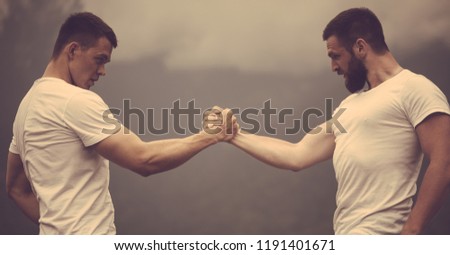 Strong male bodybuilders in white t-shirts greeting each other clasping hands together in outdoor workout over foggy mountain landscape. Rivalry, challenge, strength comparison concept