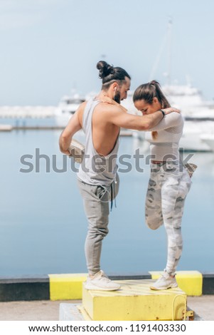 Young fitness woman and athletic man, outdoor runners stretching legs before jogging over blue sky and ocean pier background with anchored yachts.