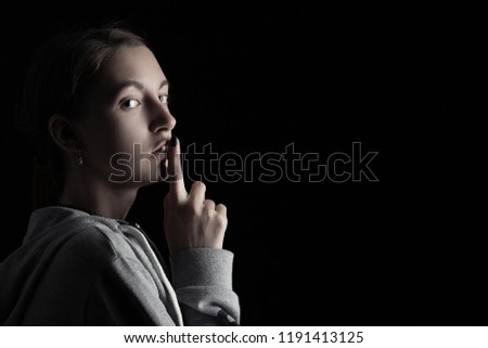 scared girl show silence gesture on black background with copy space, looking at camera