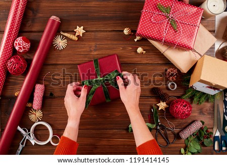 Close-up top view of female hands decorating red gift boxes with backer s twine and fir brunches on wooden table. Pine cones and christmas decoration on dark desk.