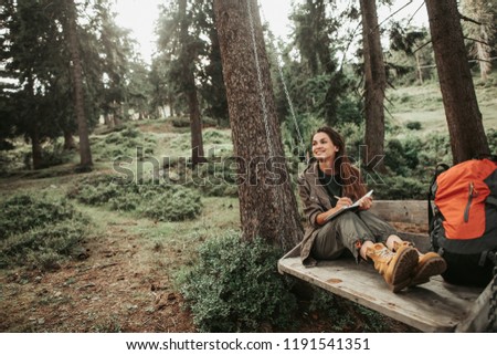 I like this place. Portrait of charming young lady holding notebook with pen while looking away with smile. Trees and green plants on background Royalty-Free Stock Photo #1191541351