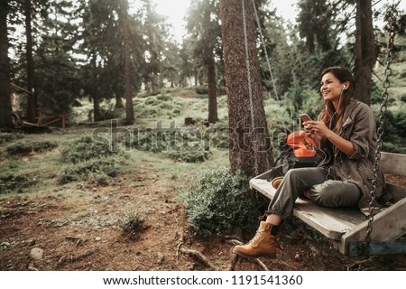 Beautiful place. Portrait of charming girl sitting on wooden swing and enjoying favorite song. She is holding mobile phone and looking away with smile Royalty-Free Stock Photo #1191541360