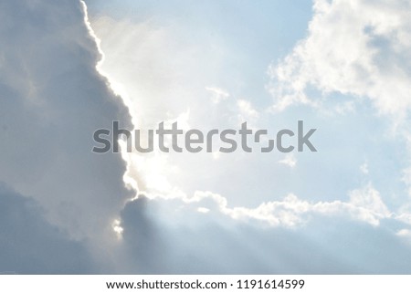 Light blue sky and puffy white and gray cloud with sun peeking out and rays streaming out.