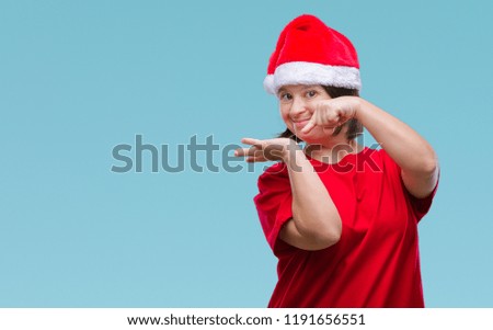 Young adult woman with down syndrome wearing christmas hat over isolated background amazed and smiling to the camera while presenting with hand and pointing with finger.