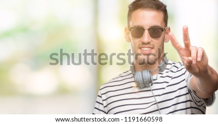 Handsome young man wearing headphones smiling looking to the camera showing fingers doing victory sign. Number two.