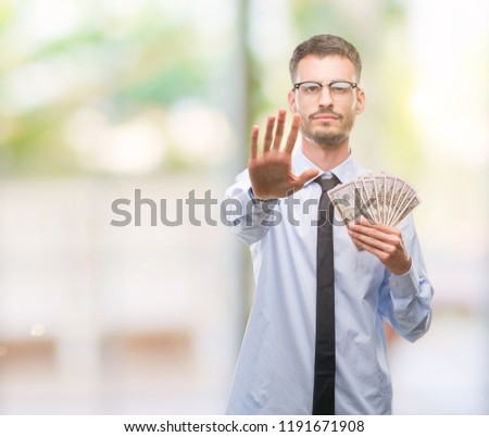 Young hipster business man holding dollars with open hand doing stop sign with serious and confident expression, defense gesture