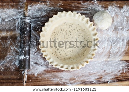 Homemade butter pie crust in pie plate with fluted pinched edge, rolling pin and extra ball of dough over floured rustic wooden background. Crust has been perforated with fork and ready for baking. Royalty-Free Stock Photo #1191681121