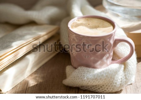 Reading a book with cup of hot cappuccino coffee. Still life with warm scarf, cup and old book on a wooden table. Winter or autumn mood concept. Warm autumn or winter picture. Selective focus. At home