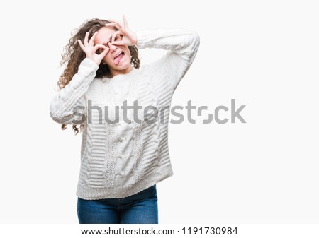Beautiful brunette curly hair young girl wearing winter sweater over isolated background doing ok gesture like binoculars sticking tongue out, eyes looking through fingers. Crazy expression.