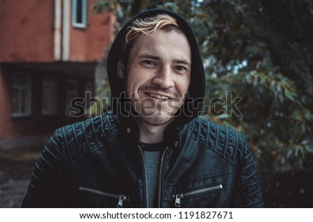 a young guy in a hood standing on the street on a cloudy day. emotional portrait of a student. street style: leather jacket and shirt