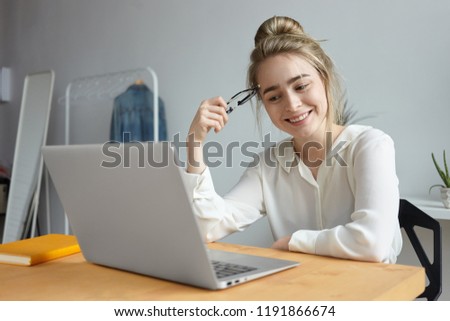 Picture of attractive cheerful student girl with hair knot surfing internet on generic portable computer, sitting at table at home, smile broadly, watching blog, communicating with friends online