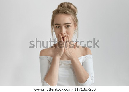 Oops! Picture of emotional beautiful young lady with hear bun covering mouth with hands, trying not to tell secret, being surprised with astonishing news or gossip, staring at camera with shocked look
