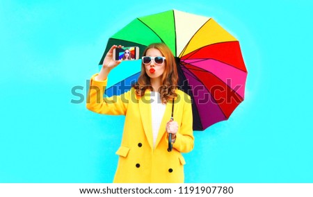 Fashion woman with colorful umbrella taking selfie by smartphone on blue wall background