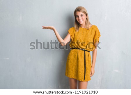 Beautiful young woman standing over grunge grey wall wearing a dress smiling cheerful presenting and pointing with palm of hand looking at the camera.