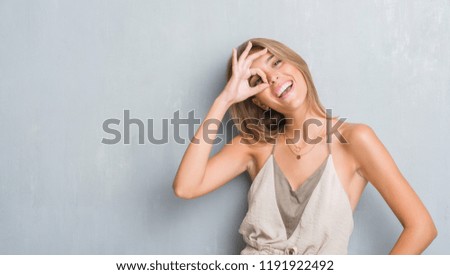 Beautiful young woman standing over grunge grey wall with happy face smiling doing ok sign with hand on eye looking through fingers