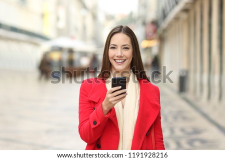 Happy woman in red looking at camera holding a phone in winter in the street
