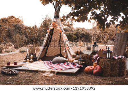 Stylish wigwam with lights, pumpkins and stack of hay outdoors. Autumn season. 