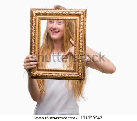 Blonde teenager woman holding vintage frame art with surprise face pointing finger to himself