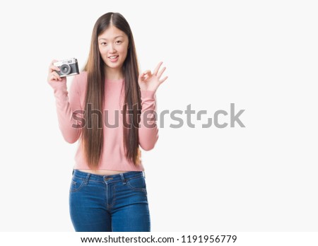 Young Chinese woman over isolated background taking pictures using vintage camera doing ok sign with fingers, excellent symbol