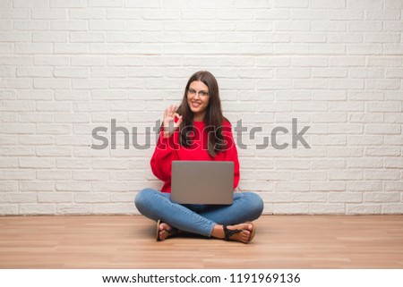 Young brunette woman sitting on the floor over white brick wall using laptop doing ok sign with fingers, excellent symbol
