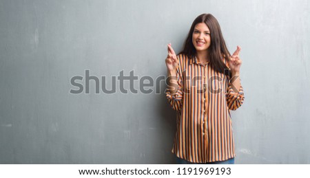 Young brunette woman over grunge grey wall smiling crossing fingers with hope and eyes closed. Luck and superstitious concept.