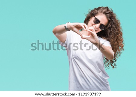 Beautiful brunette curly hair young girl wearing sunglasses over isolated background smiling in love showing heart symbol and shape with hands. Romantic concept.