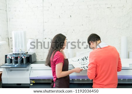 Rear view of busy printing specialists examining banner with text and standing at large format printer in office