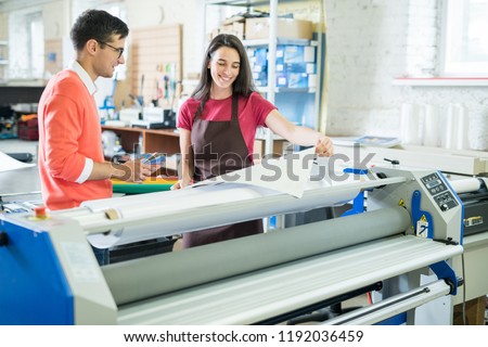 Cheerful excited beautiful lady in apron showing printed banner to manager who picking out color on swatch for printing