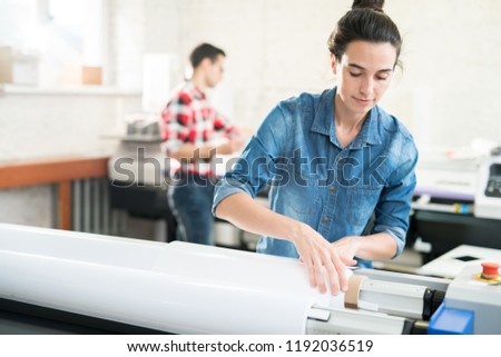 Serious confident young woman  with hair bun loading wide format printer while working at printing house Royalty-Free Stock Photo #1192036519