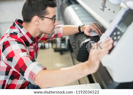 Serious thoughtful handsome young engineer in shirt examining wide format printer while holding inspection in printing house Royalty-Free Stock Photo #1192036522