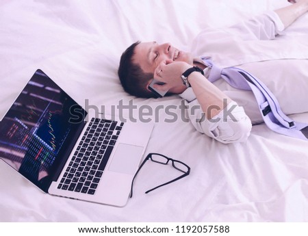 Young happy trader businessman is lying on a white bed in a room and talking on the phone.