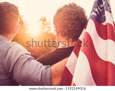 Two cute guys holding the US flag against the backdrop of the rays of the beautiful setting sun. View from the back. Preparing for the holidays