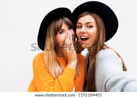 Friendship, happiness and people concept. Two smiling girls whispering gossip on white background.  Orange sweater, black similar hats. 