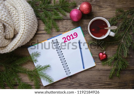 Beautiful Christmas picture layout postcard with 2019 new year: a mug of tea, a notebook with a pen for plans, fir branches with toys, a warm scarf on a wooden rustic background, top view