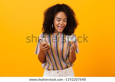 Picture of excited happy african woman posing isolated over yellow background using mobile phone holding credit card.