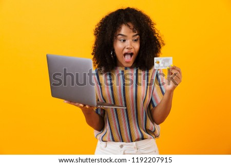 Photo of excited shocked african woman posing isolated over yellow background using laptop computer holding credit card.