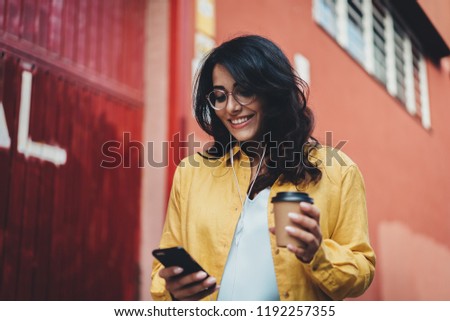 Young positive woman in optical eyeglasses smiling while typing an sms message on smartphone device, attractive hispanic student girl listening to music and reading new on cellphone outdoors