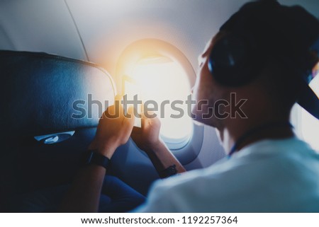 Young hipster guy wearing cap sitting in airplane and using modern smartphone and listening to music in headphones, male traveller typing sms message while sitting in aircraft, flare light