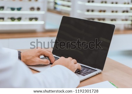 partial view of optician using laptop with blank screen in optica Royalty-Free Stock Photo #1192301728