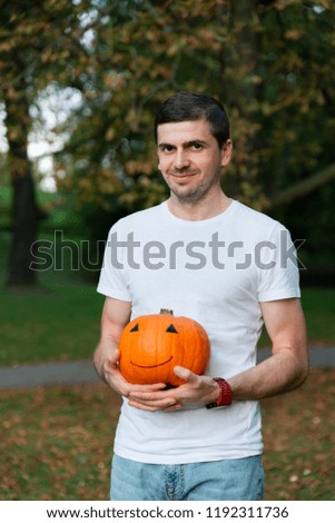 Handsome brunette man in white t-shirt holding orange pumpkin with drawing happy face and posing at camera. Concept of preparing  for Halloween party and autumn holidays.