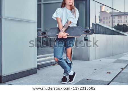 Cropped photo of a young hipster girl dressed in white shirt and ripped jeans holds skateboard while posing near skyscraper.