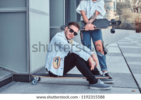 Trendy dressed couple - young hipster man sitting on a longboard and his girlfriend standing near outdoors.
