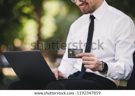Cropped photo of handsome business man outdoors in the park using laptop computer holding credit card.